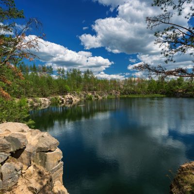 A forest lake with rocky shores and coniferous trees, with a blue sky and white clouds reflected in the water