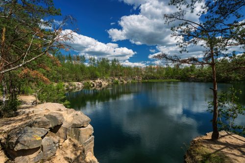 A forest lake with rocky shores and coniferous trees, with a blue sky and white clouds reflected in the water